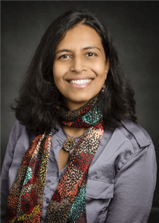 Vidya Madhavan mit dunklem Haar, Schal und breitem Lächeln, vor dunklem Hintergrund.